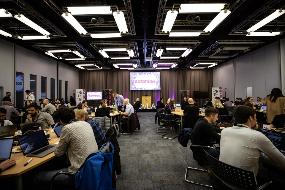 People sitting at tables in a digital theatre with a large screen showing Botathon